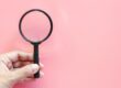 A hand holding a black magnifying glass in front of a light pink background representing how to find an egg donor