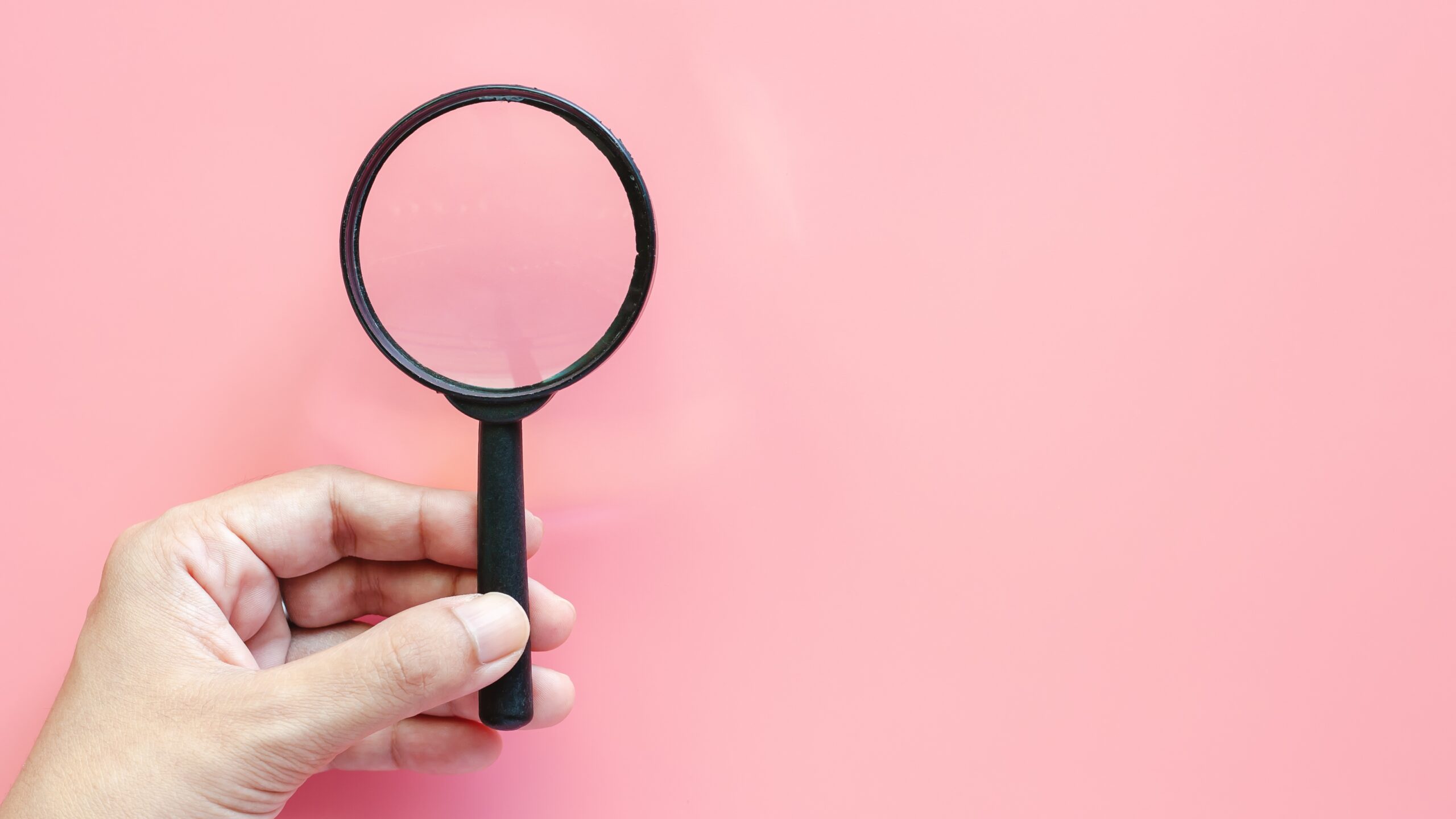 A hand holding a black magnifying glass in front of a light pink background representing how to find an egg donor