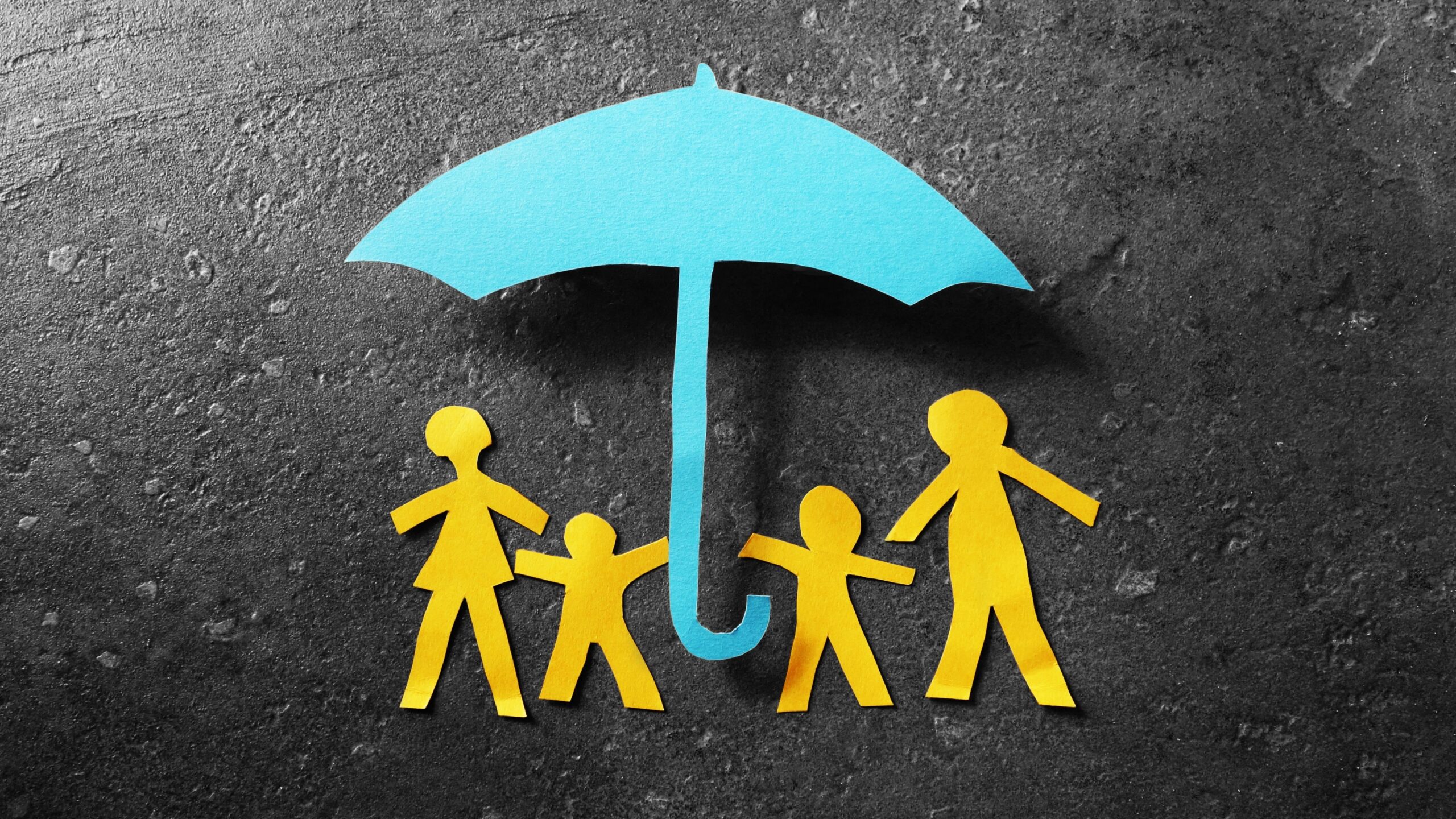 Yellow cut out paper family of 4 under a light blue paper cut out umbrella laid against a dark concrete background. Representing the question, does insurance cover donor eggs?