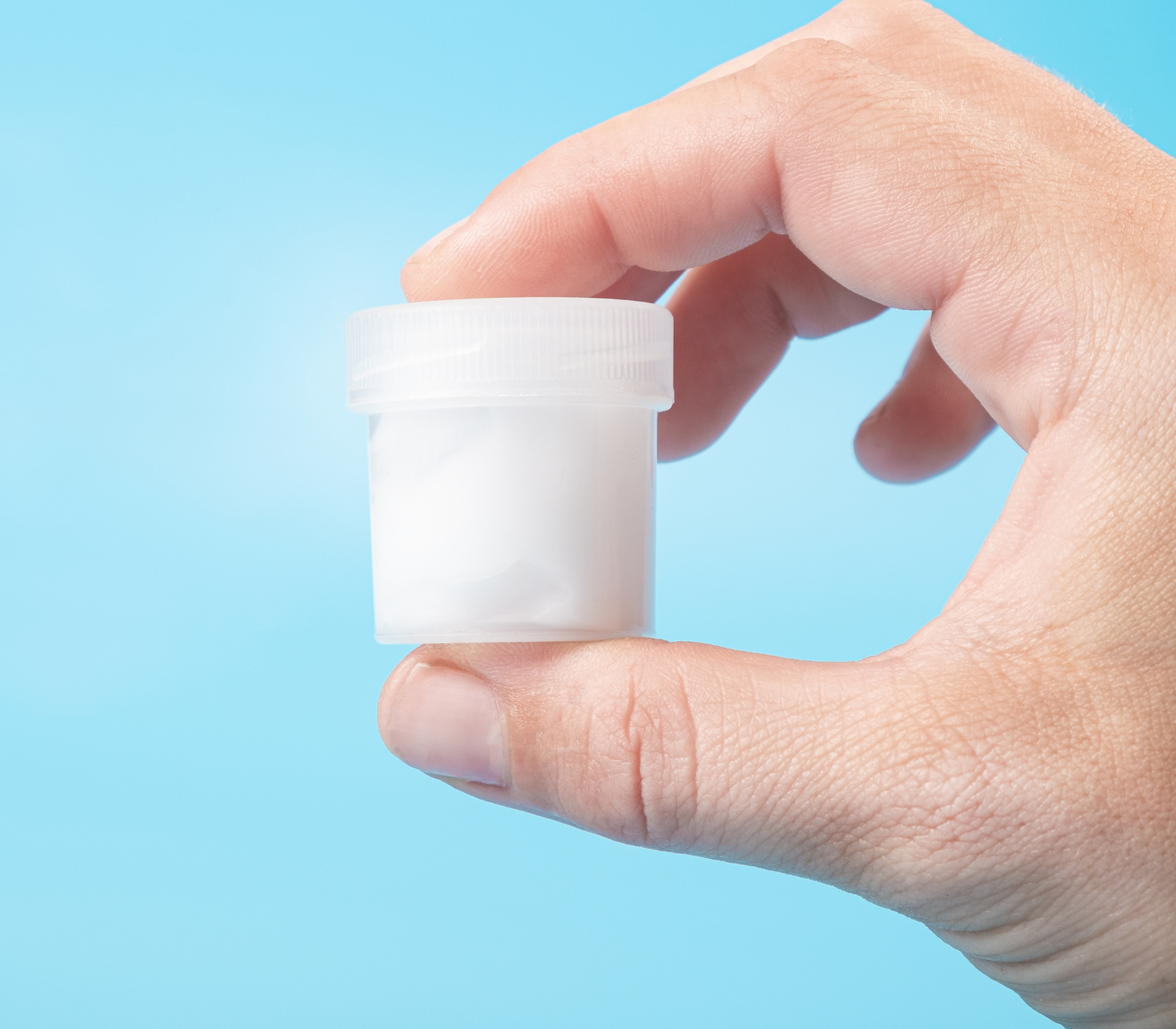 A mans hand holding a sperm donation cup in front of a light blue background.