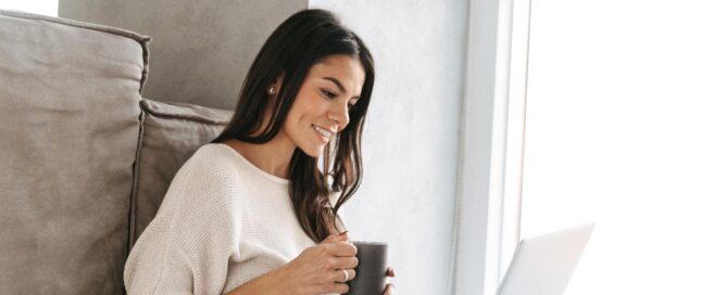 Women sitting on the floor with a cup of coffee, on her computer, smiling.