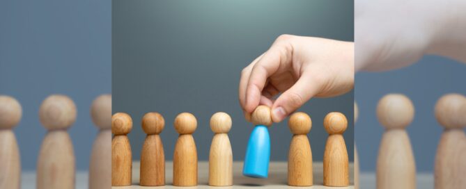 An image of wooden shaped people with a hand reaching down and grabbing one of the wooden dolls representing how old do you have to be to donate sperm.